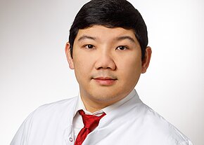 Frontalporträt eines Mannes in weißem Kittel und roter Krawatte vor hellem Hintergrund; ruhiger, direkter Blick.