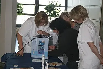 Vier Frauen, darunter eine Krankenschwester, stehen um einen Tisch herum und schauen interessiert auf ein Informationsmaterial. Sie scheinen sich über ein Thema auszutauschen und diskutieren gemeinsame Anliegen. Pentagramm im Hintergrund.