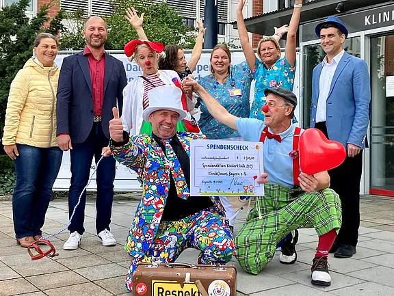 Spende für die Klinik Clowns Vor einem Klinikum feiern mehrere Erwachsene; zwei Klinikclowns präsentieren einen großen Spendenscheck, zeigen Daumen hoch. Ein rotes Herz und bunte Kostüme fallen auf. Die Gruppe lächelt und jubelt; Anlass ist eine Spende zugunsten Klinikclown-Einsätze im Krankenhaus.