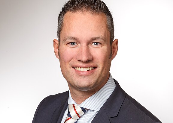 Kopf-und-Schulter-Porträt einer lächelnden Person im Business-Outfit vor neutralem Hintergrund; kurzes dunkelgraues Haar, helles Hemd, dunkler Anzug und gestreifte Krawatte; Blick direkt in die Kamera.