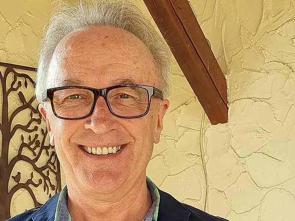 Nahaufnahme eines lächelnden Mannes mit grauem Haar und dunkler Brille. Er schaut direkt in die Kamera; sein Gesicht ist gut ausgeleuchtet. Neutraler, heller Hintergrund.
