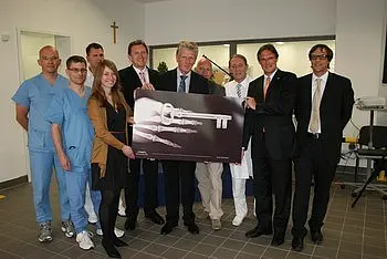 Eine Gruppe von Personen steht zusammen, die einen großen symbolischen Schlüssel präsentiert. In lässiger Kleidung sind medizinisches Personal und weitere Beteiligte zu sehen, die sich über den Schlüssel freuen. Die Szenerie vermittelt einen feierlichen Anlass, möglicherweise die Eröffnung einer neuen Einrichtung.