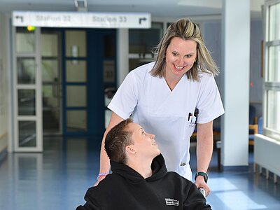 Eine Krankenschwester lächelt und spricht lächelnd mit einem jungen Mann in einem Rollstuhl. Der Hintergrund zeigt einen hellen Krankenhausflur mit Türschildern und Sitzmöglichkeiten. Die beiden wirken freundlich und kommunikativ.