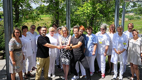 Gruppe von Pflegekräften und Mitarbeitenden steht lächelnd im Freien. Zwei Personen vorn überreichen sich eine Trophäe oder Auszeichnung, die Gruppe schaut zu. Stimmung festlich, Sonnenschein, Bäume im Hintergrund.