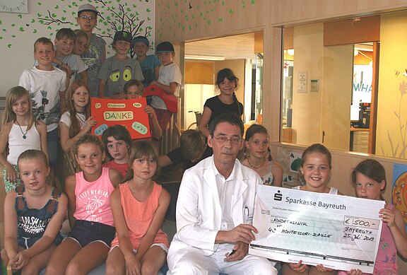 Gruppe von Kindern in einer Klinik überreicht gemeinsam mit einem Arzt einen symbolischen Spendenscheck. Ein Mädchen hält den großen Scheck der Sparkasse, andere Kinder zeigen ein Schild mit „Danke“. Fröhliche Atmosphäre auf einer Kinderstation.