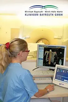 Eine Mitarbeiterin im Krankenhaus sitzt vor einem Computer, auf dem ein Röntgenbild des Skeletts angezeigt wird. Sie trägt einen blauen Kittel und hat eine Brille auf. Im Hintergrund ist ein medizinisches Gerät sichtbar. Oben im Bild ist das Logo der Klinik zu sehen.
