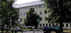Eine mehrstöckige, gelb gestrichene Gebäudefront mit Fenstern. Vor dem Gebäude stehen Bäume und Autos. Der Himmel ist klar und es ist tagsüber.