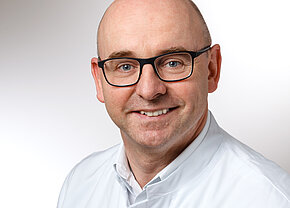 Dr. med. Andreas Zumbrägel Ein lächelnder Mann mit Glatze und Brille trägt einen weißen Arztkittel. Sein Gesicht ist gut beleuchtet, und er schaut direkt in die Kamera. Der Hintergrund ist neutral und hell gehalten.