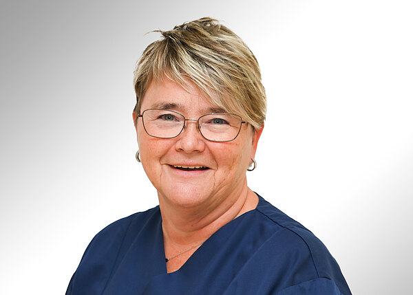Porträt einer lächelnden Frau mit kurzem blond-braunem Haar und Brille, in dunkelblauer Berufskleidung vor hellem Hintergrund.