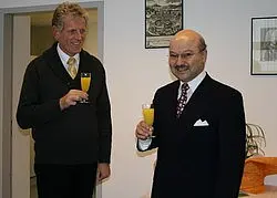 Zwei Männer stehen in einem Raum und stoßen mit Gläsern an. Der Mann links trägt einen grauen Pullover und sieht lächelnd aus. Der Mann rechts ist elegant gekleidet und hat einen Bart. Beide wirken freundlich und erfreut, während sie einen orangefarbenen Drink in der Hand halten.