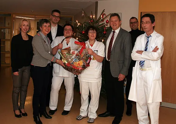 Mehrere Klinikmitarbeitende und Besucher stehen in einem Flur; zwei Pflegekräfte halten einen großen Geschenkkorb. Im Hintergrund ein geschmückter Weihnachtsbaum. Die Gruppe lächelt; es wirkt wie eine festliche Übergabe.