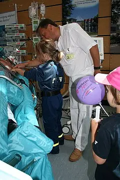 In einem Krankenhausraum untersucht ein Arzt einen Patienten, während ein Kind in einer blauen Uniform und ein weiteres Kind mit einem Ballon daneben stehen. Die Szenerie vermittelt eine freundliche und lehrreiche Atmosphäre im medizinischen Umfeld.