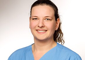 Porträt einer lächelnden Frau in einem hellblauen medizinischen Kittel. Ihr Haar ist halblang und leicht gewellt, sie trägt kleine Perlenohrringe. Der Hintergrund ist hell und schlicht, was den Fokus auf ihr freundliches Gesicht lenkt.