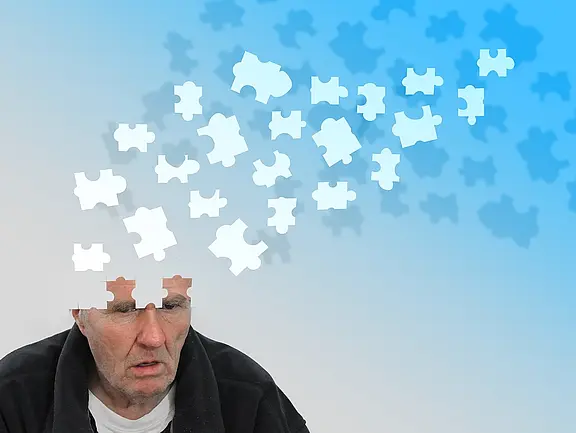 Ein älterer Mann schaut nachdenklich und traurig, während Puzzlestücke aus seinem Kopf schweben. Der Hintergrund ist hellblau und grau, was eine melancholische Atmosphäre schafft. Die verschwindenden Puzzlestücke symbolisieren vergessene Gedanken oder verlorenes Wissen.