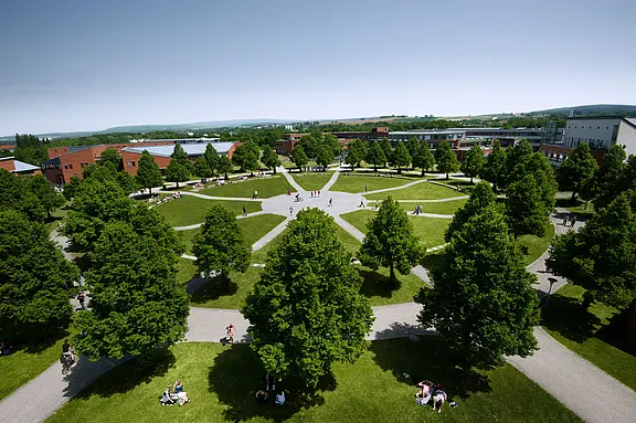 Großer, kreisförmig angelegter Campuspark mit Rasenflächen, vielen Bäumen und radialen Wegen, die sich in der Mitte treffen. Zahlreiche Studierende sitzen oder gehen im Sonnenschein. Um den Park stehen moderne Hochschulgebäude.
