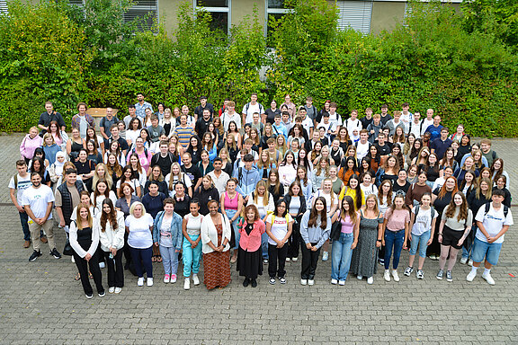 Gruppenfoto von etwa 150 Schülern und Schülerinnen, die auf einem Schulhof stehen. Sie lächeln und sind in verschiedenen Kleidung gewählt. Im Hintergrund sind grüne Pflanzen und ein Gebäude sichtbar. Es herrscht eine freundliche und ausgelassene Atmosphäre.