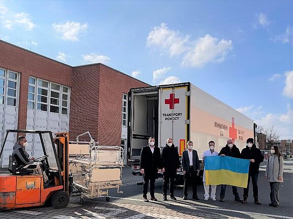 Vor einem Gebäude wird ein Lkw mit Rotem-Kreuz-Logo beladen. Ein Gabelstapler hebt Krankenhausbetten in den Auflieger. Daneben steht eine Gruppe, einige mit Masken, Personen halten die ukrainische Flagge. Szene zeigt einen Hilfstransport.