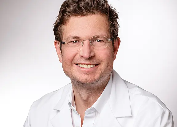 Porträt eines lächelnden Mannes mit Brille in weißem Kittel vor hellem, neutralem Hintergrund; er blickt direkt in die Kamera.
