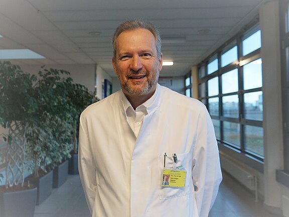 Mann mittleren Alters im weißen Kittel mit Ausweis lächelt in einem hellen Krankenhausflur. Fokus auf einen freundlichen medizinischen Mitarbeiter.