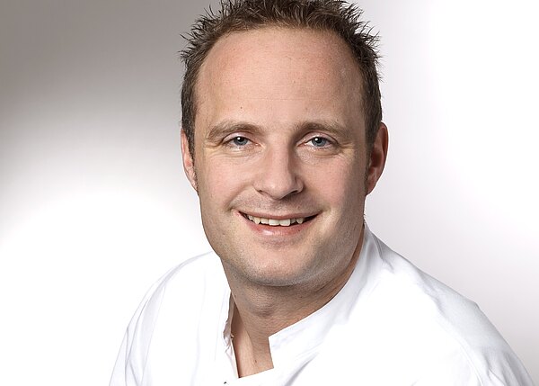 Porträtfoto eines lächelnden Mannes mit kurzen Haaren in weißer Berufskleidung, der direkt in die Kamera schaut, vor neutral hellem Hintergrund.