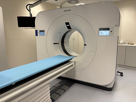 Moderner Computertomograph in einem Klinikraum: ein großer, ringförmiger Scanner mit integrierten Bedienanzeigen und herausgefahrenem Patiententisch mit blauer Auflage, bereit für eine Untersuchung.