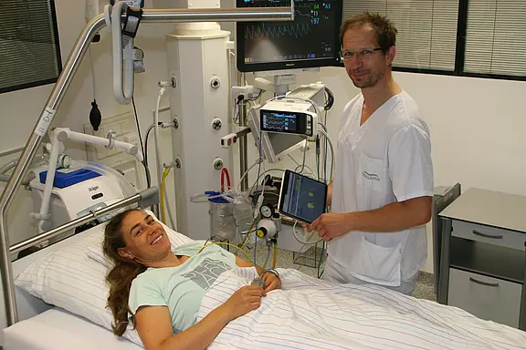 In einem Krankenhauszimmer liegt eine lächelnde Patientin im Bett. Daneben steht medizinisches Personal in weißer Kleidung und überwacht mit Tablet und Monitoren die Vitalwerte. Die angeschlossenen Geräte deuten auf eine laufende Untersuchung oder Behandlung.