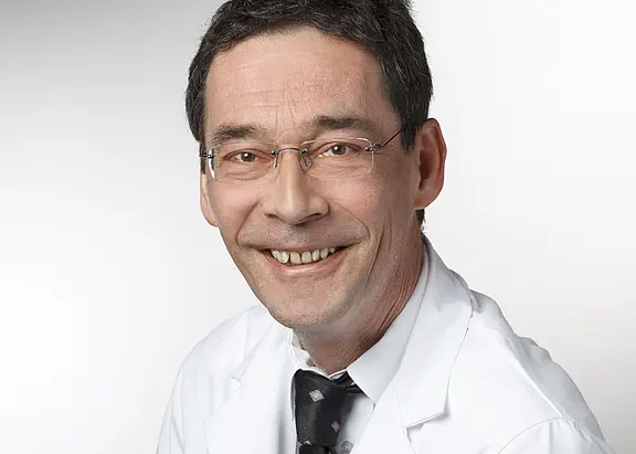 Porträt eines lächelnden Mannes mit Brille im weißen Arztkittel und Krawatte vor hellem, neutralem Hintergrund; blickt direkt in die Kamera.