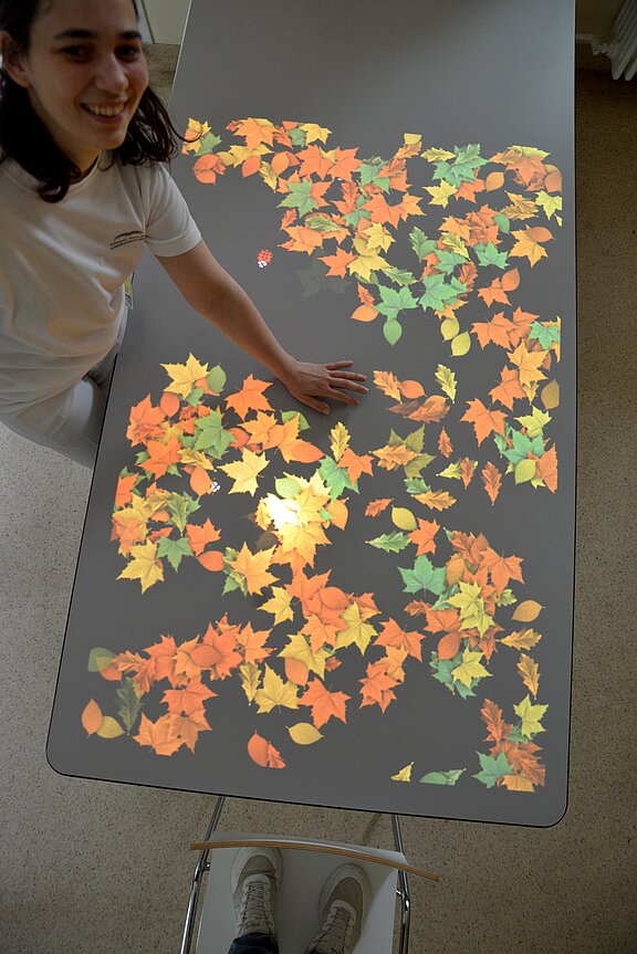 Eine Person steht an einem Tisch, auf dem leuchtend farbige Herbstblätter projiziert sind. Die Blätter in Gelb, Orange und Grün sind unregelmäßig verteilt, während die Person mit einer Hand darüber fährt und lächelt. Der Tisch hat eine einfarbige Oberfläche.