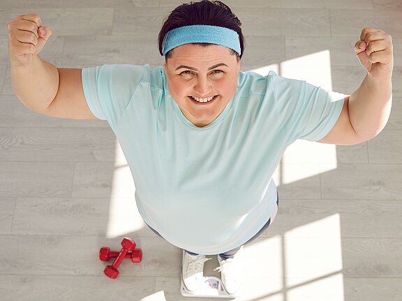 Fröhliche Person im Sportoutfit steht auf einer Badezimmerwaage und reckt beide Fäuste triumphierend; Blick nach oben zur Kamera. Daneben liegen kleine Hanteln auf dem Boden.