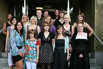 Eine Gruppe von 15 Personen steht lächelnd auf einer Treppe vor einem Gebäude. Die Frauen tragen verschiedene Outfits, von casual bis elegant. Es herrscht eine fröhliche Stimmung, und alle scheinen sich zu amüsieren.