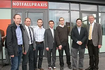 Sechs Männer stehen lächelnd vor einem Eingangsbereich mit der Aufschrift „Notfallpraxis“. Sie tragen unterschiedliche formelle Kleidung und wirken in einer freundlichen, einladenden Pose. Der Hintergrund zeigt moderne Architektur des Gebäudes.