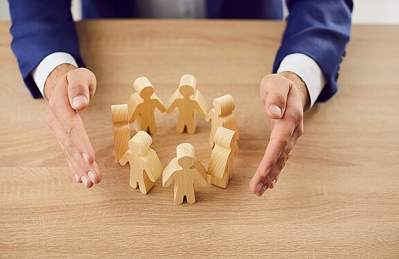 Ein paar Holzfiguren stehen händehaltend in einem Kreis auf einem Tisch. Die Hände eines Mannes, der einen Anzug trägt, umrahmen die Figuren schützend, was Gemeinschaft und Schutz symbolisiert.