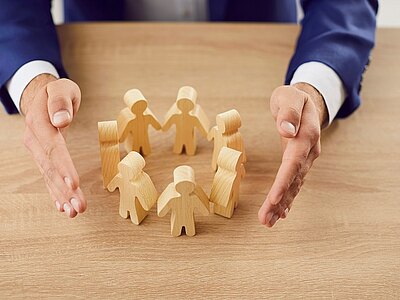 Ein paar Holzfiguren stehen händehaltend in einem Kreis auf einem Tisch. Die Hände eines Mannes, der einen Anzug trägt, umrahmen die Figuren schützend, was Gemeinschaft und Schutz symbolisiert.