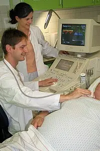 Zwei Ärzte arbeiten gemeinsam an einem Ultraschallgerät. Der männliche Arzt bedient das Gerät und untersucht einen Patienten, der auf einer Liege liegt. Eine weibliche Ärztin beobachtet die Untersuchung. Das Umfeld wirkt klinisch und professionell.