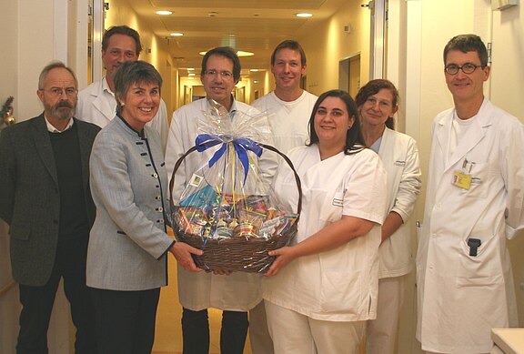 Gruppe von Krankenhausmitarbeitern steht in einem Klinikflur. Eine Frau überreicht gemeinsam mit einer Pflegekraft einen großen Geschenkkorb mit blauer Schleife. Alle lächeln, als Zeichen des Dankes und der Anerkennung.