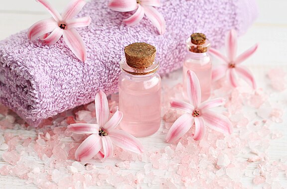 Wellness-Szene: Zusammengerolltes lila Handtuch, zwei kleine Glasfläschchen mit rosafarbenem Öl oder Duftwasser, umgeben von rosa Badesalz und zarten Blüten. Symbolisiert Entspannung und Spa-Atmosphäre.