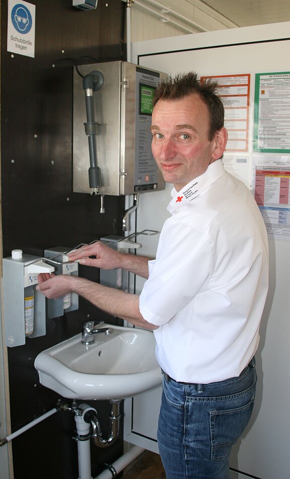 Ein Mann in einem weißen Hemd steht an einem Waschbecken und drückt mit einer Hand auf einen Seifenspender. Hinter ihm ist eine Wand mit verschiedenen Geräten und Sicherheitshinweisen. Der Mann lächelt und scheint bereit, sich die Hände zu waschen.