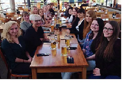 Eine Gruppe von zwanzig Personen sitzt an einem langen Tisch in einem Restaurant. Vor ihnen stehen Gläser mit Bier. Die Atmosphäre wirkt gesellig und einladend. Die Frauen und Männer lächeln und scheinen an einem gemeinsamen Ereignis oder Treffen teilzunehmen.