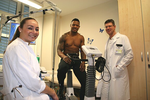 In einem Klinikraum absolviert ein oberkörperfreier Patient auf einem Ergometer einen medizinischen Belastungstest. Zwei medizinische Fachkräfte in weißen Kitteln stehen daneben, lächeln und überwachen die Untersuchung.