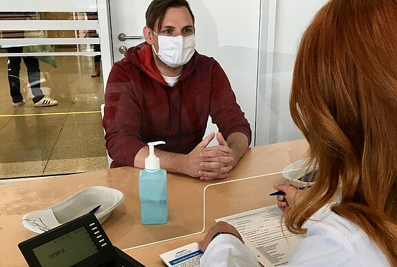 Ein Mann mit Schutzmaske sitzt hinter Plexiglas einem medizinischen Mitarbeitenden gegenüber. Die Fachkraft füllt ein Formular aus; auf dem Tisch steht Desinfektionsmittel. Szene einer medizinischen Anmeldung oder Beratung, vermutlich im Zusammenhang mit Impfung oder Test.