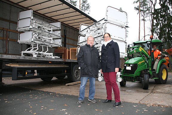 Zwei Personen stehen vor einem offenen Lkw, auf dem mehrere gestapelte Krankenhausbetten geladen sind; daneben ein grüner Traktor mit Fahrer. Szene einer Anlieferung beziehungsweise eines Transports medizinischer Betten im Freien.