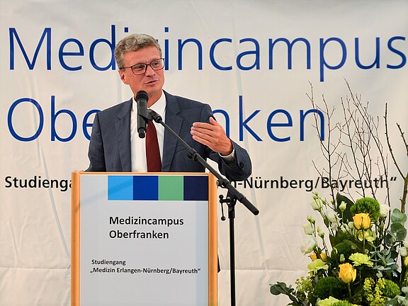 Ein Redner steht an einem Mikrofon und spricht zu einem Publikum. Im Hintergrund ist ein Banner mit der Aufschrift „Medizincampus Oberfranken“ zu sehen. Auf einem Tisch vor ihm befinden sich Blumenarrangements.