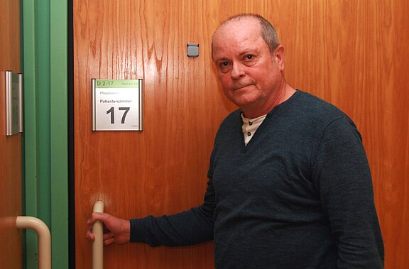 Ein älterer Mann steht vor einer Holztür und hält den Türgriff. An der Tür hängt ein Schild mit der Aufschrift „Patientenzimmer 17“. Er blickt ruhig in die Kamera.