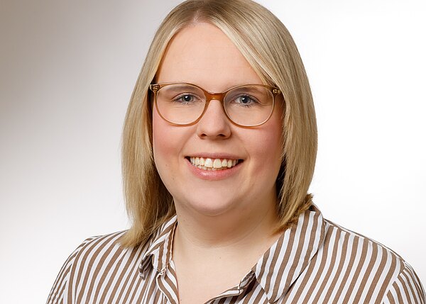 Nahaufnahme eines professionellen Porträts: lächelnde Person mit schulterlangen blonden Haaren und goldbrauner Brille, trägt ein braun-weiß gestreiftes Hemd vor hellem, neutralem Hintergrund.