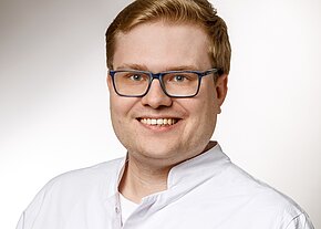 Porträt eines freundlich lächelnden Erwachsenen mit blonden Haaren und dunkelblauer Brille, in weißem Kittel vor neutralem Hintergrund.