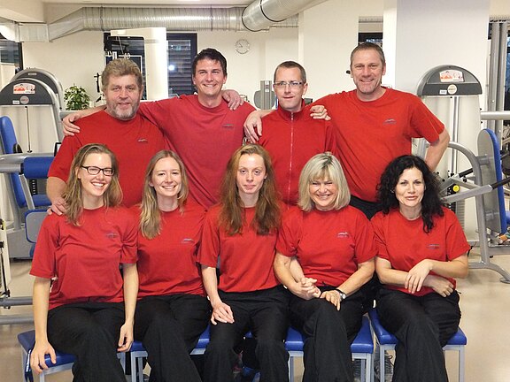 Neun Personen in einheitlichen roten T-Shirts posieren in einem Fitnessstudio; fünf sitzen vorn, vier stehen dahinter, alle lächeln.
