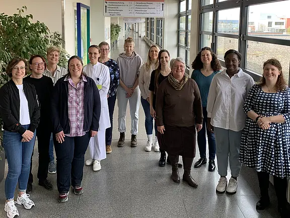 Gruppe von etwa 14 Menschen steht in einem hellen Flur vor großen Fenstern und schaut lächelnd in die Kamera. Unterschiedliche Kleidung, darunter eine Person im weißen Kittel. Innenraum mit Tageslicht; freundliche, gemeinschaftliche Atmosphäre.
