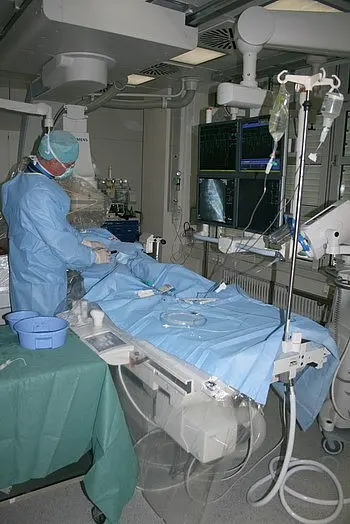 Ein Arzt in Schutzkleidung steht neben einem OP-Tisch, der mit blauer Abdeckung versehen ist. An der Wand sind Monitoranwendungen sichtbar, die medizinische Daten anzeigen. Der Raum wirkt steril und ist mit medizinischen Geräten ausgestattet.