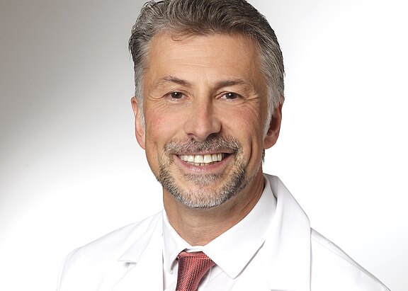 Ein lächelnder Arzt trägt einen weißen Kittel und eine dunkle Krawatte. Der Hintergrund ist hell und neutral. Das Bild vermittelt Professionalität und Freundlichkeit.