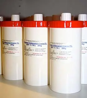 Fünf weiße Behälter mit roten Deckeln stehen nebeneinander. Jeder Behälter enthält 200 Gramm einer Hautcreme, die zur Behandlung von speziellen Hautzuständen verwendet wird. Die Etiketten auf den Behältern geben den Produktnamen und die Inhaltsstoffe an.